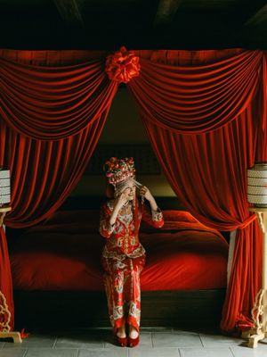 Bride in traditional Chinese red wedding dress standing before ornate red canopy bed.