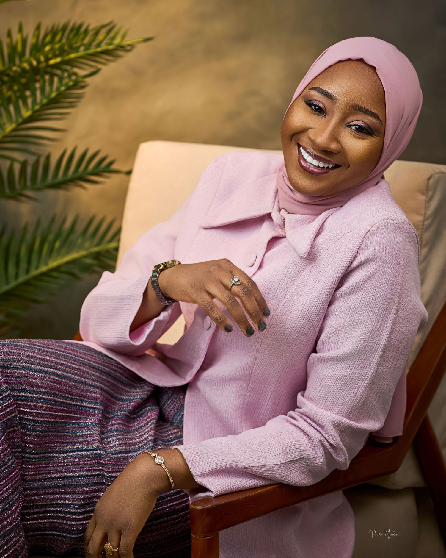 Portrait of a woman in pink hijab and blazer smiling warmly in an indoor setting.