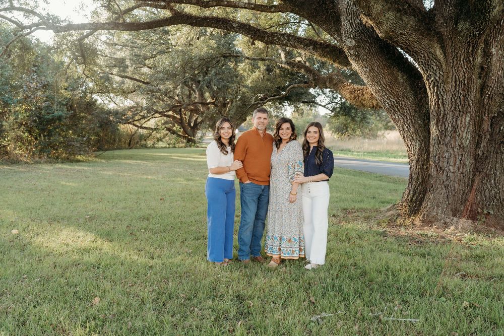 A group of people stand together under large oak trees in a grassy field during golden hour.