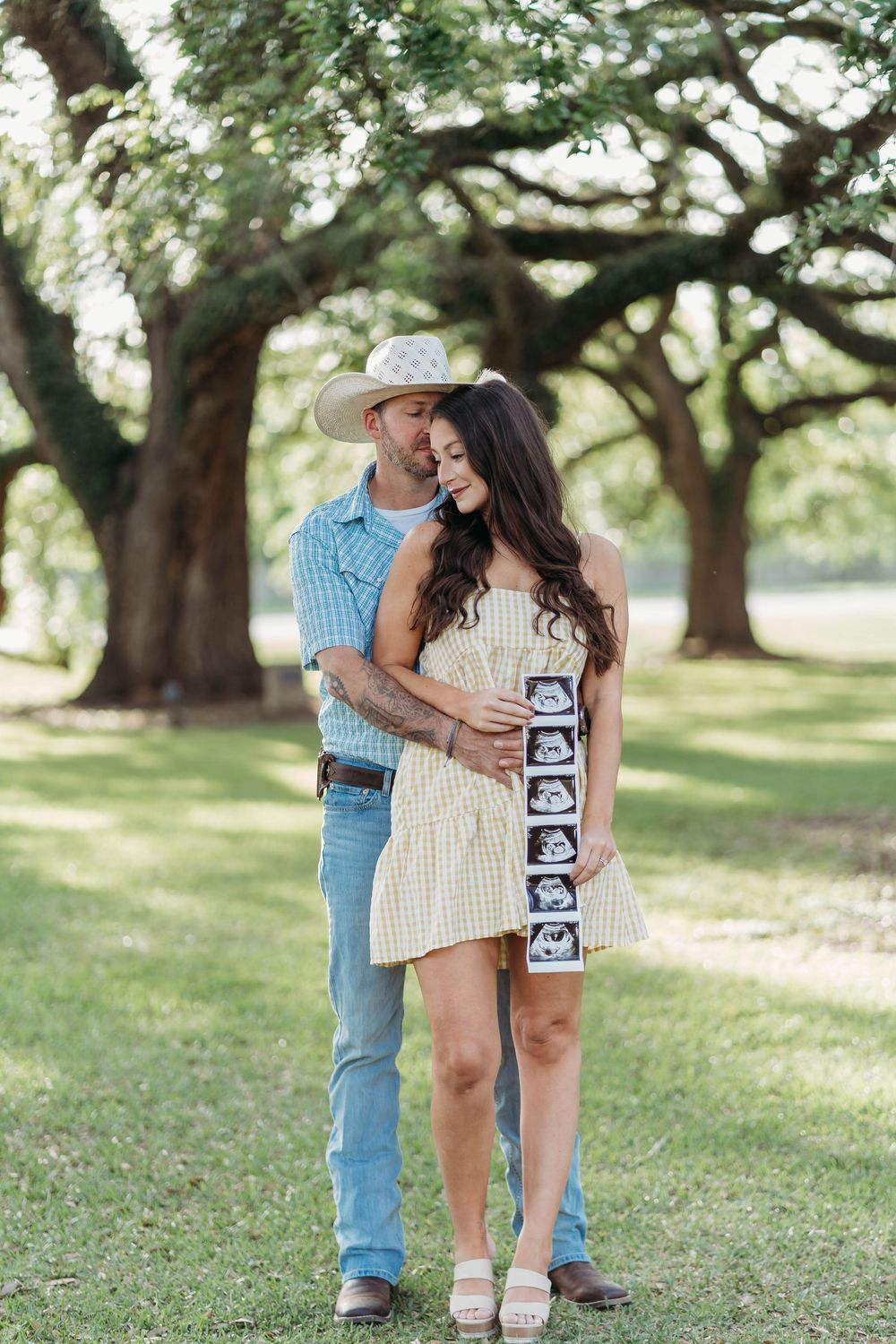 Couple embracing outdoors holding ultrasound photos, pregnancy announcement in a park with large oak trees.
