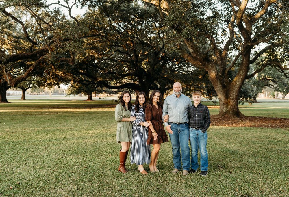 People standing together beneath sprawling tree branches in a sunlit outdoor setting.