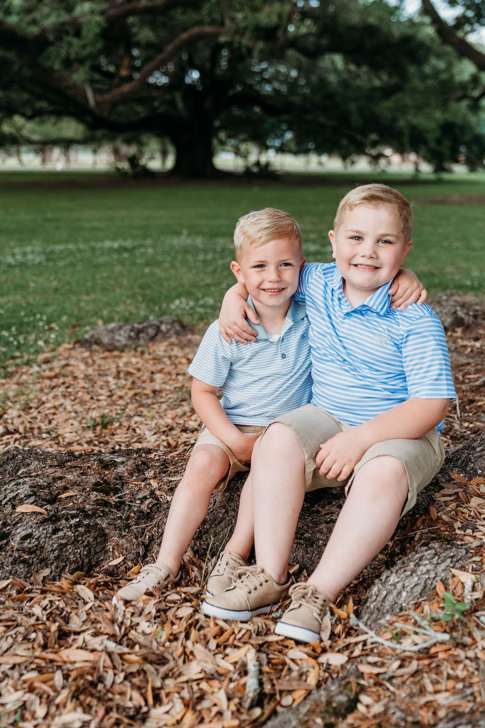Two smiling boys in matching blue polo shirts and khaki shorts sit together on tree roots in a park.