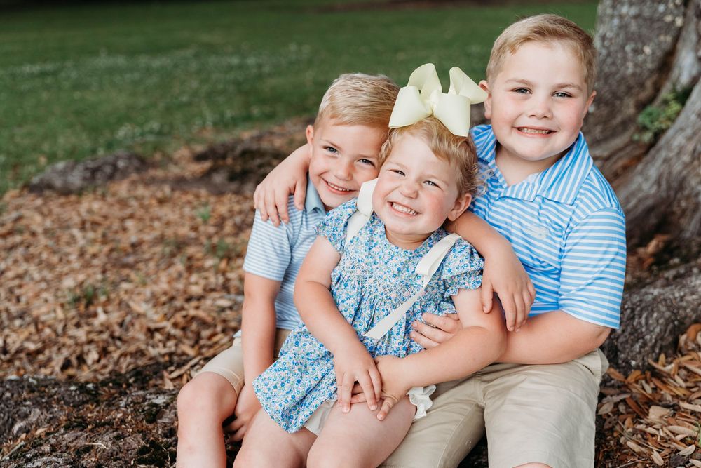 Three smiling children pose together outdoors, wearing coordinating blue outfits, sitting near a tree.
