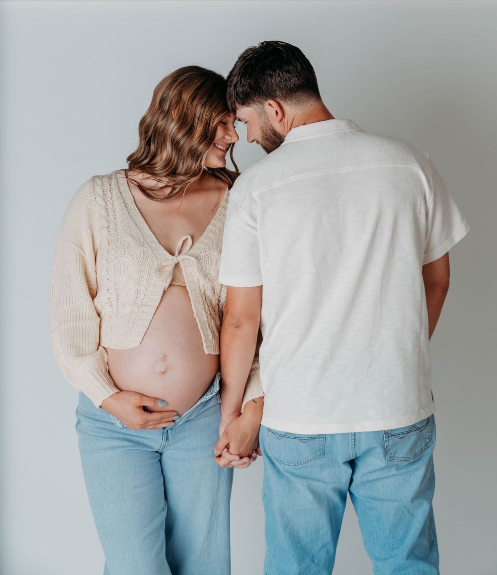 Pregnant couple holding hands in studio, woman in cream cardigan showing baby bump, man in white shirt, both in jeans.