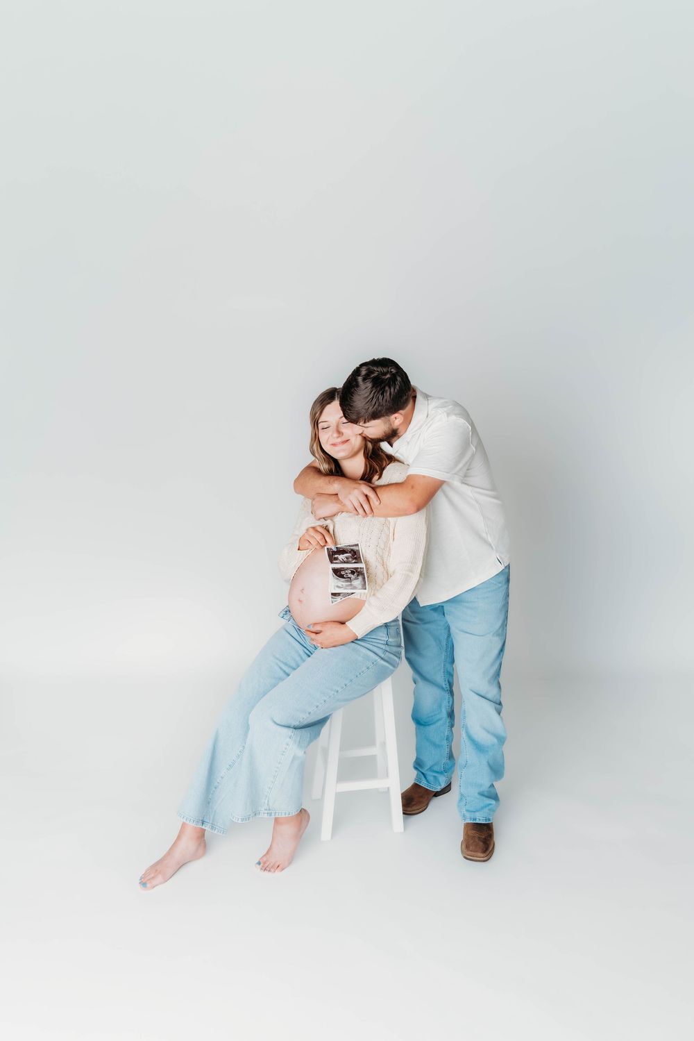 Pregnant woman sitting on stool holding ultrasound photo while partner embraces her from behind on grey background.