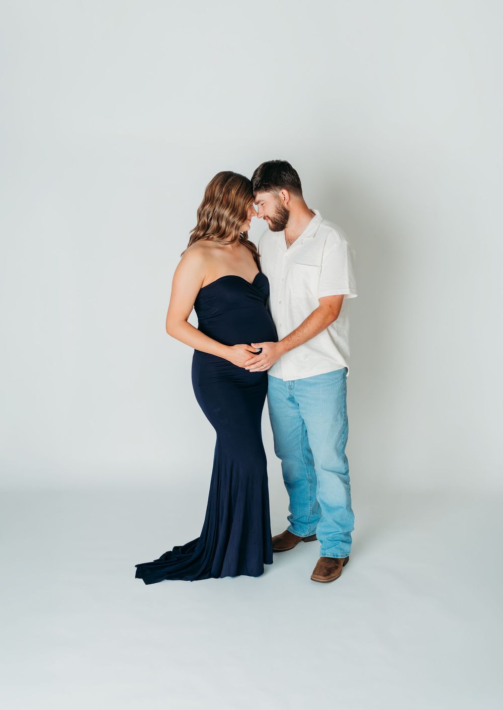 Pregnant couple in maternity photo, woman in navy gown, man in white shirt and jeans, tenderly touching foreheads in studio.