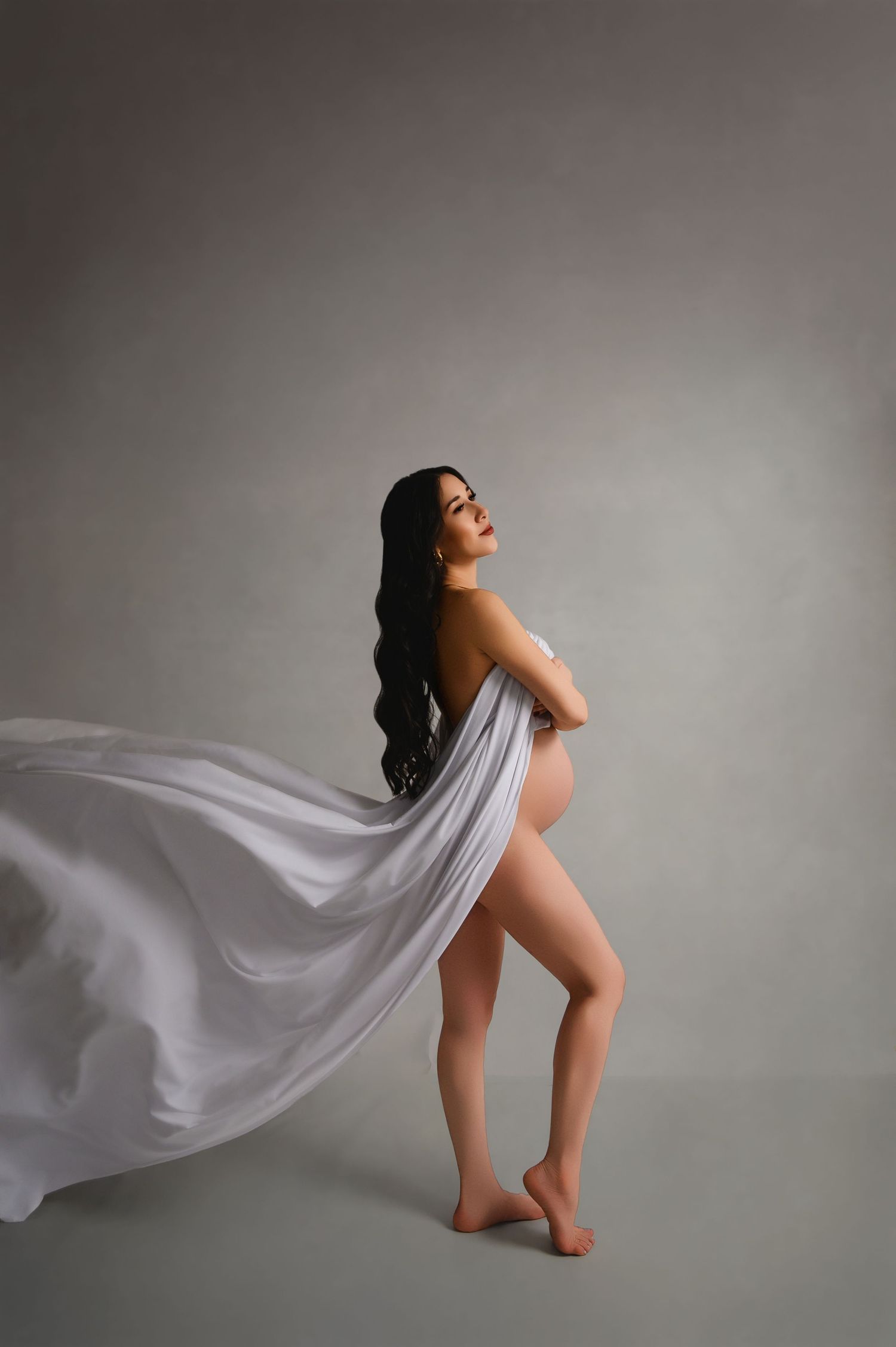 Artistic maternity portrait with flowing white fabric against grey studio background.