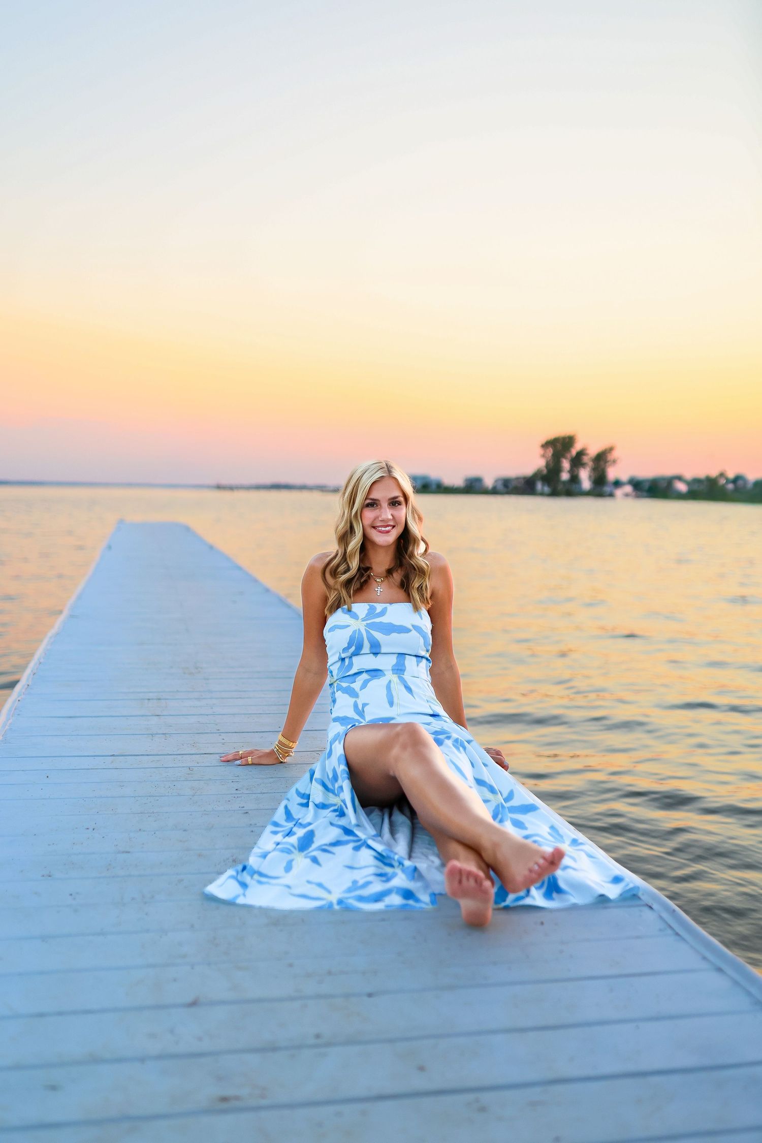 Blonde woman in blue floral strapless dress sitting on white dock at colorful sunset.