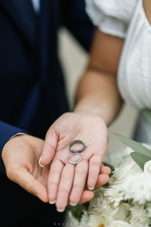 Two silver wedding rings rest in an open palm next to white flowers, capturing an intimate wedding detail shot.