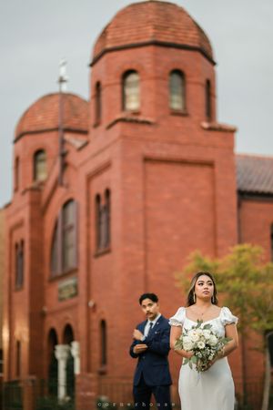 Wedding couple poses in front of a historic red brick church with twin domed towers on an autumn day.