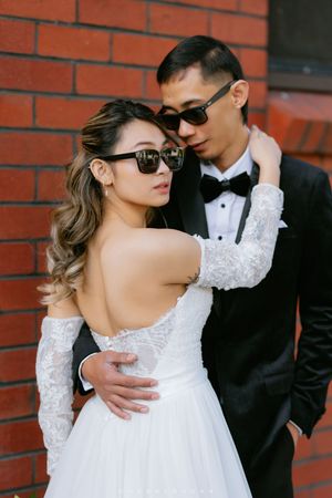 Stylish couple in wedding attire and sunglasses pose against red brick wall at outdoor photoshoot.