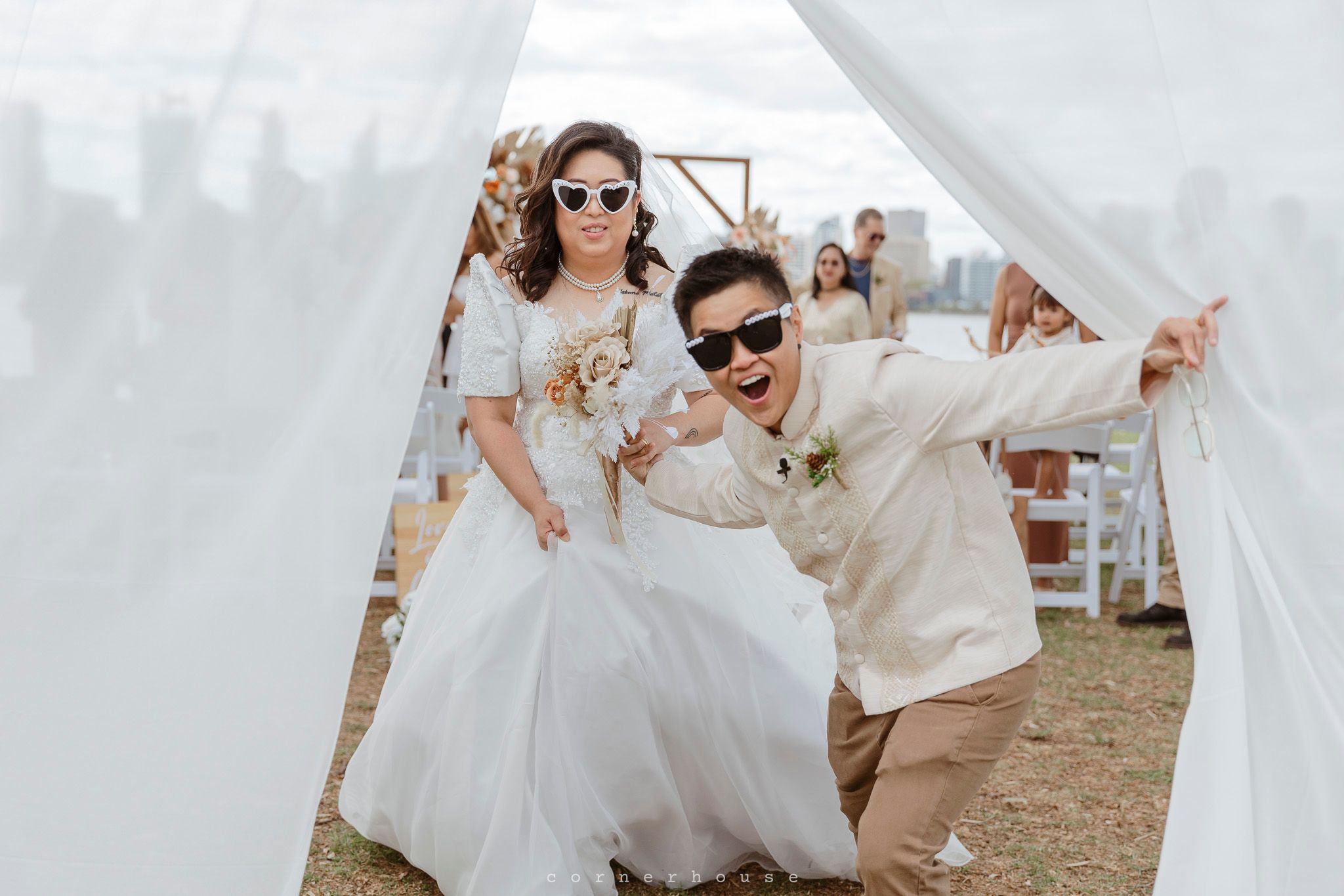 A joyful wedding couple wearing sunglasses pose playfully under a white tent at an outdoor celebration.