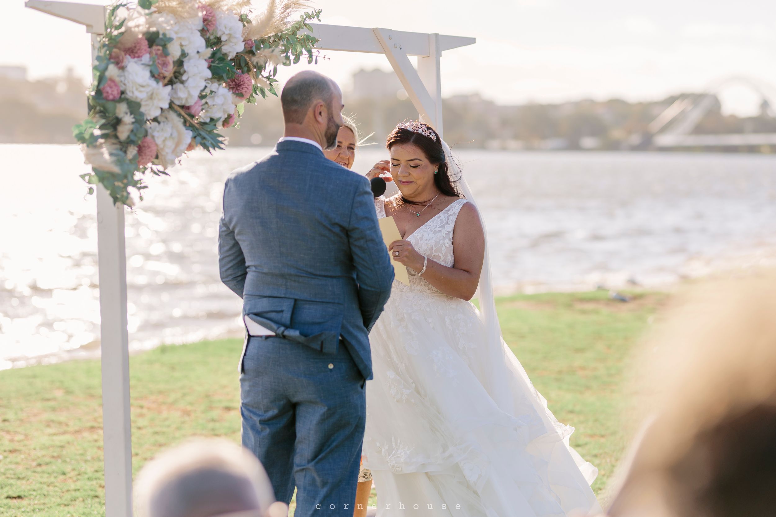 Couple exchanges vows under a floral arch during waterfront outdoor wedding ceremony at sunset.