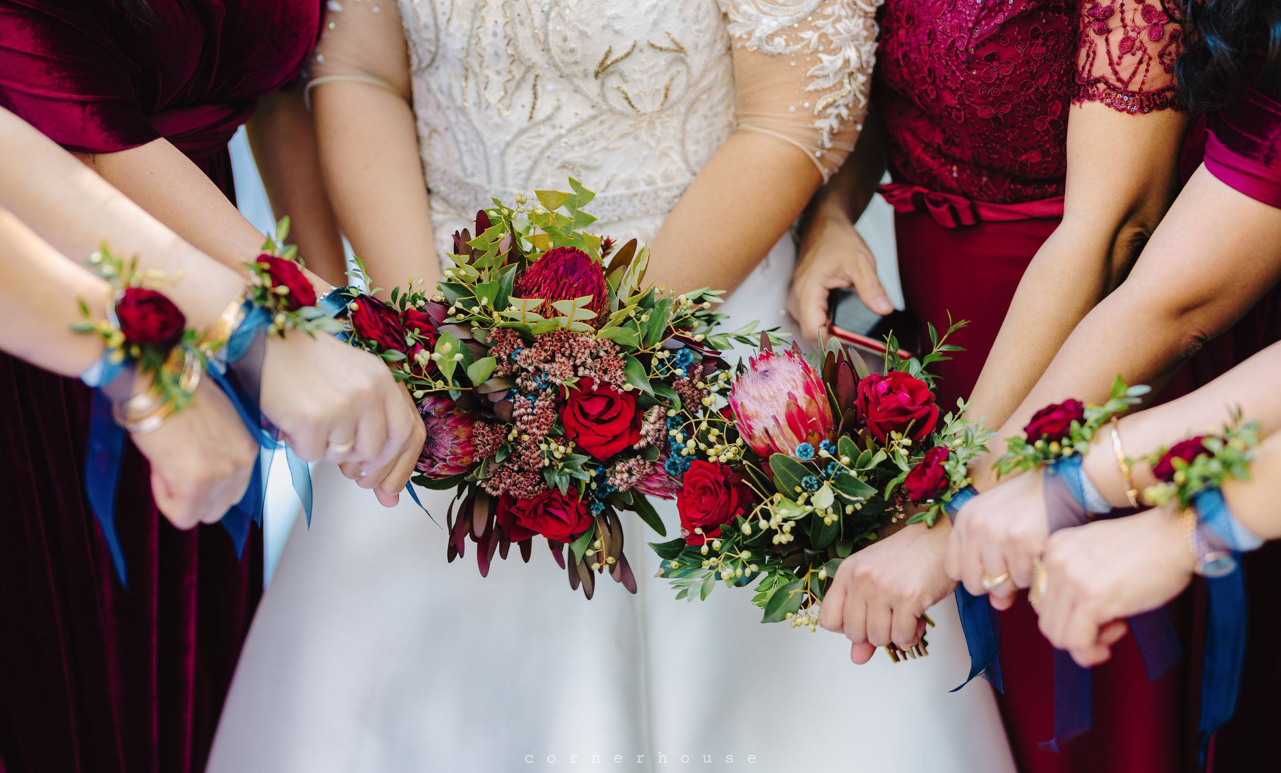 Arms of bride and bridesmaids wearing velvet dresses display colorful floral corsages and bouquets with red roses.