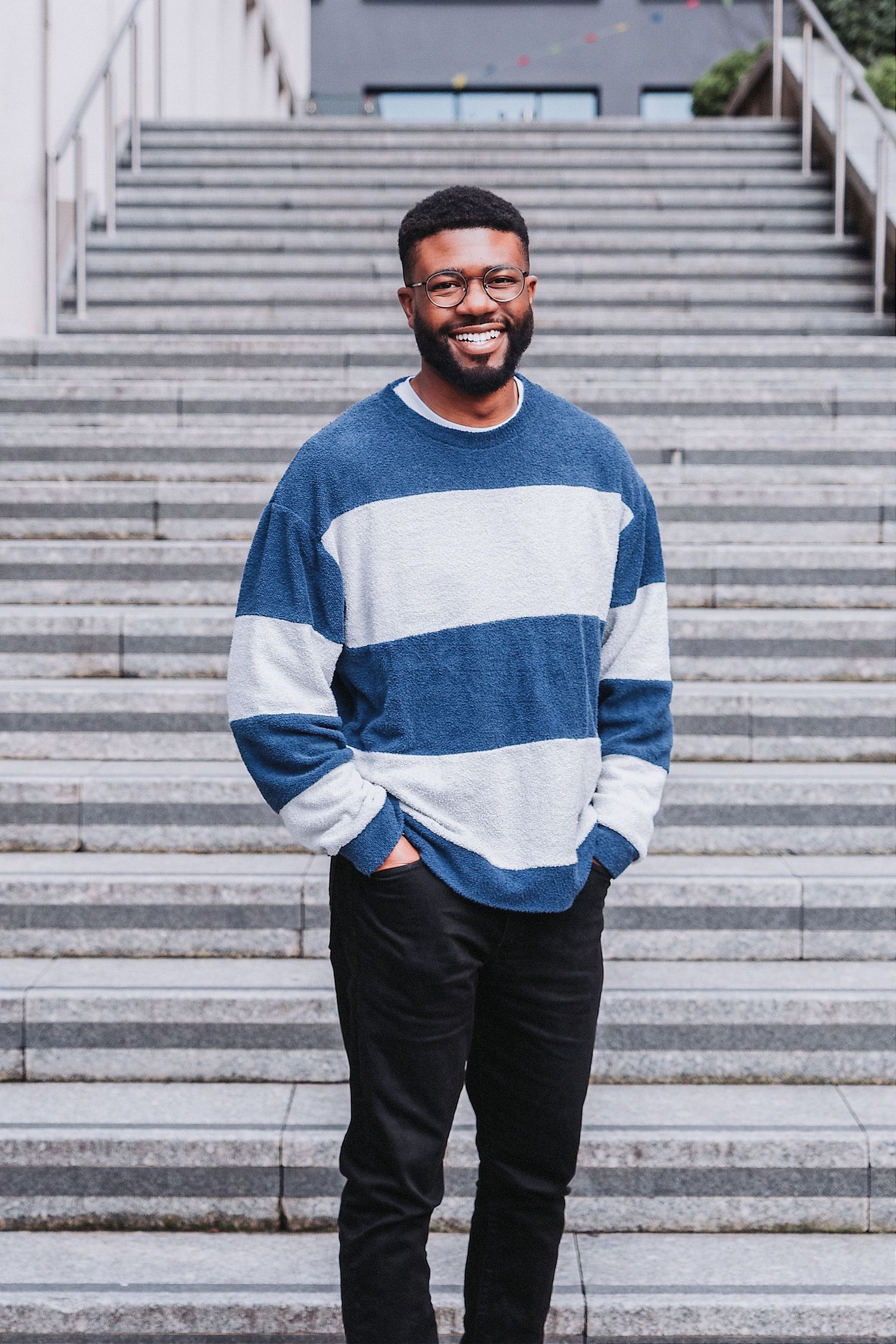 A person in a blue and white striped sweater stands smiling on stone steps in an outdoor setting.