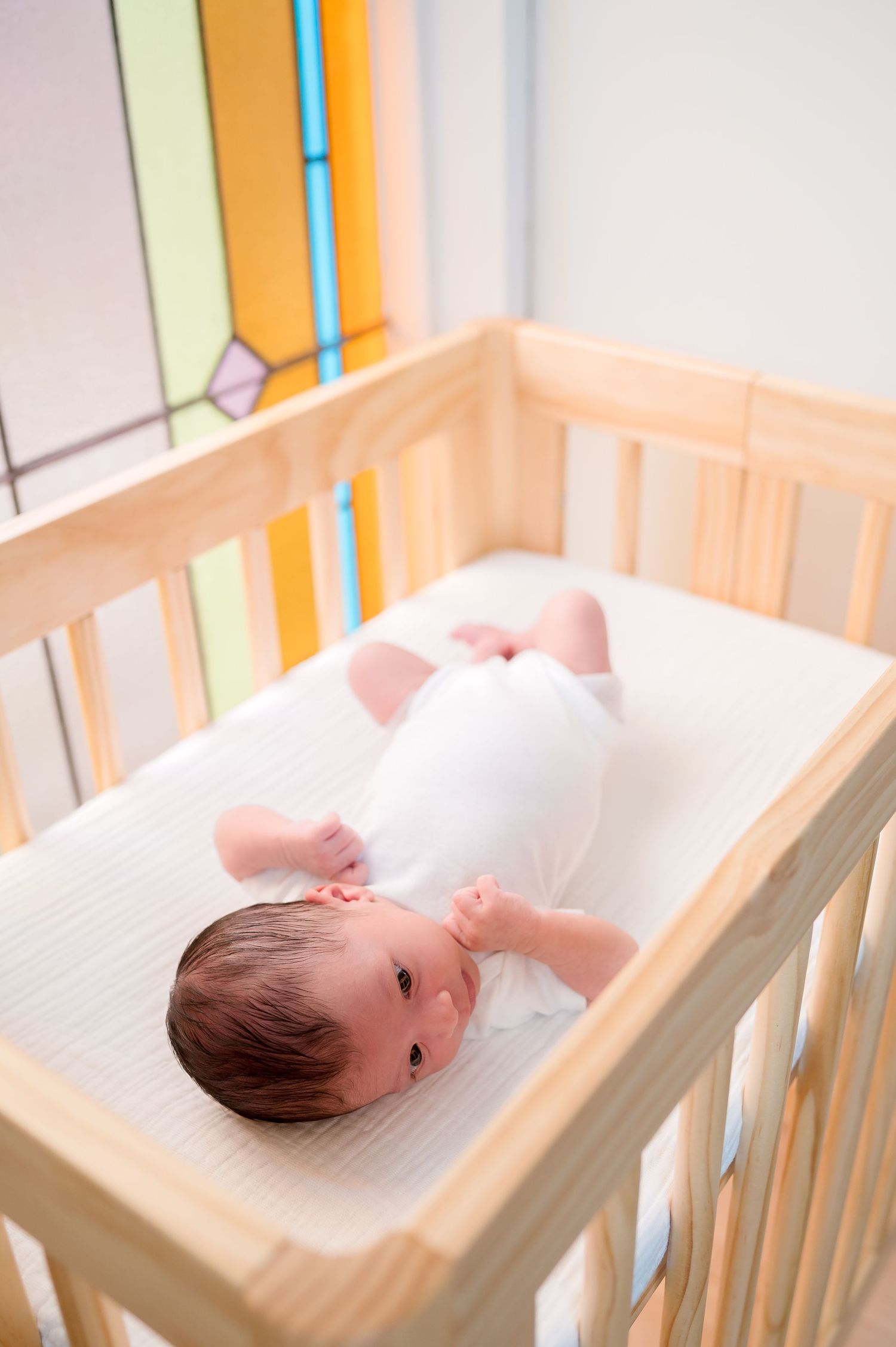 A baby in white sleepwear lies on their back in a wooden crib with light blue bars.