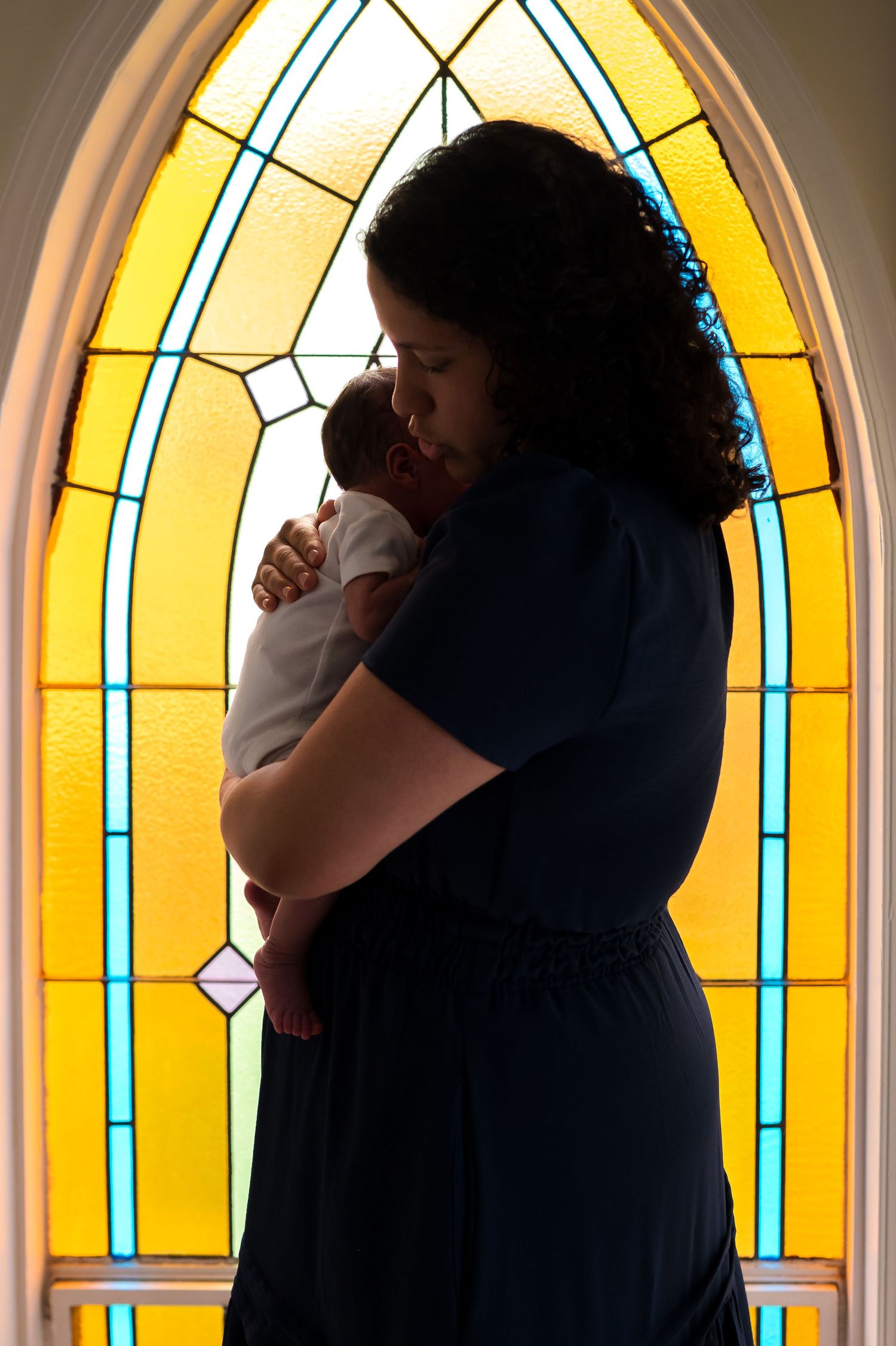 Silhouetted figures embrace in front of a bright stained glass church window with yellow and blue geometric patterns.