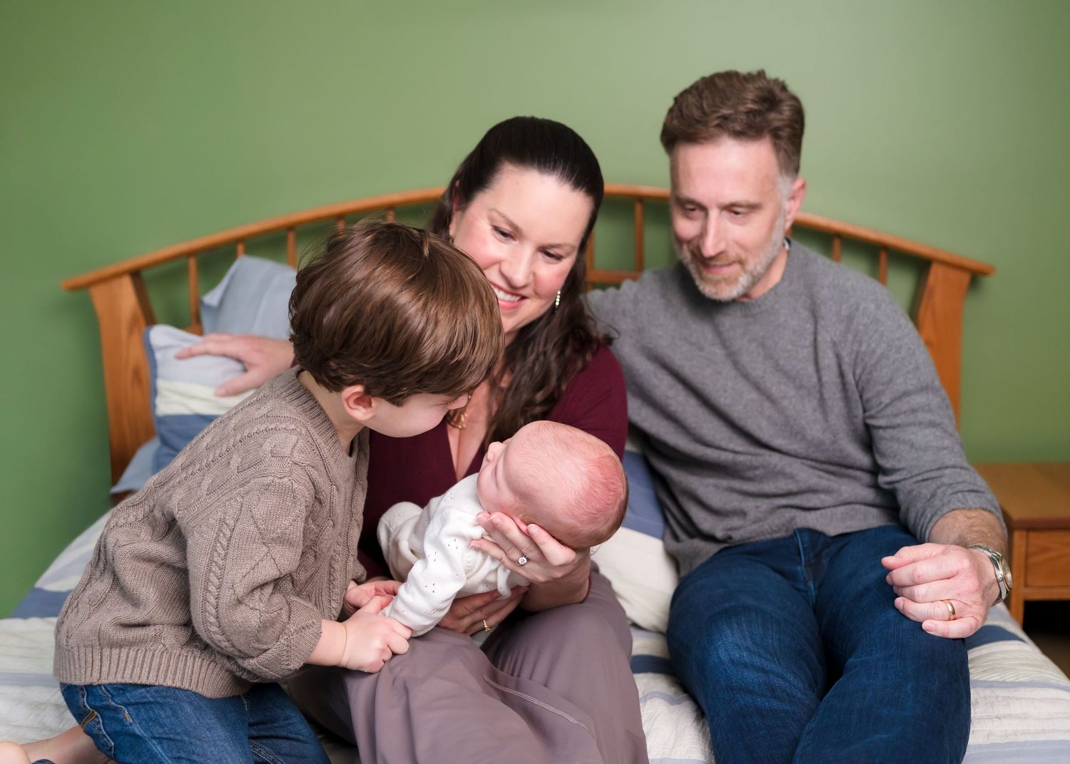 Family of four sitting on bed, parents smiling at newborn baby held by older child in cozy home setting.