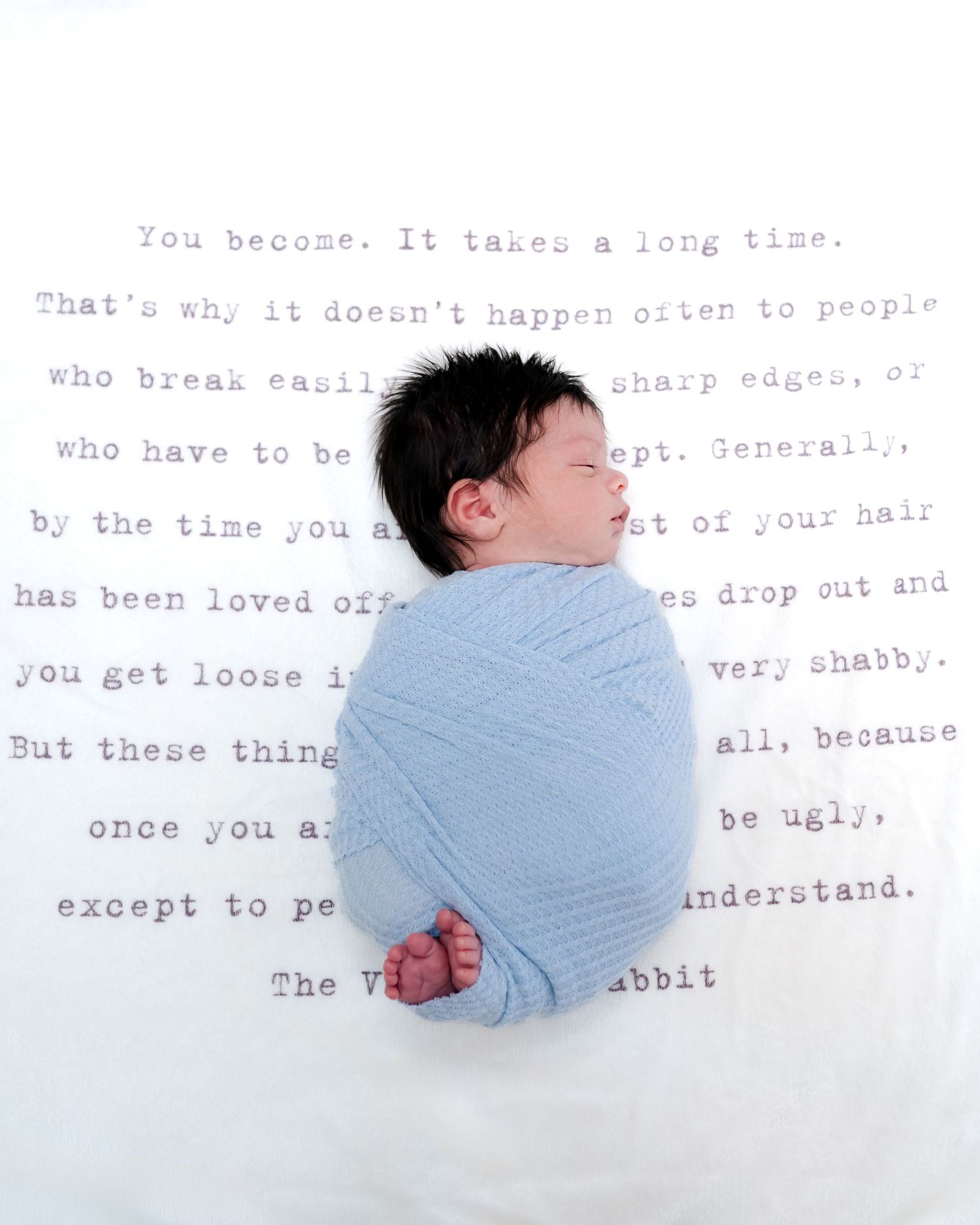 Newborn baby swaddled in blue wrap lying on white blanket printed with Velveteen Rabbit quote.