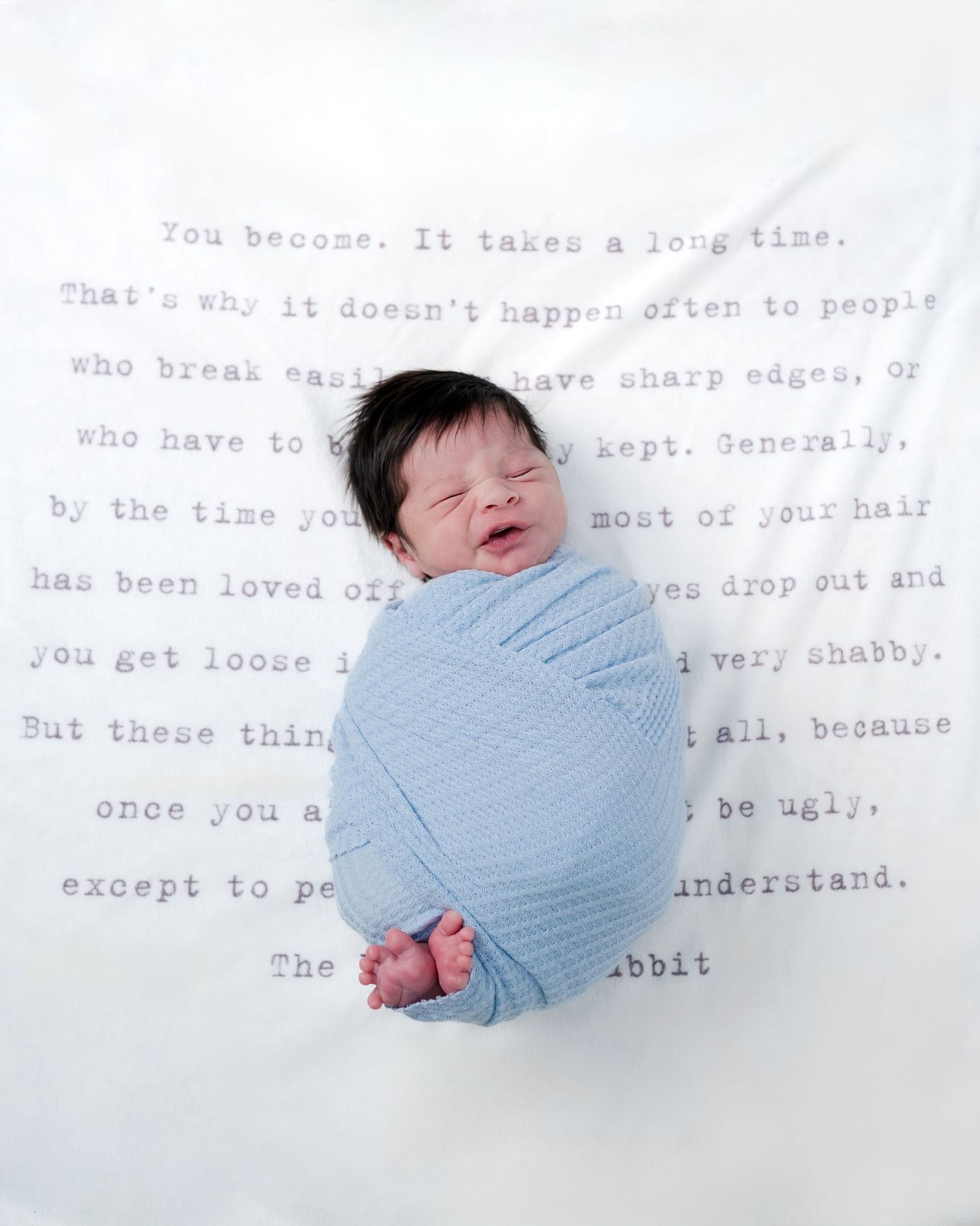 Newborn baby swaddled in blue wrap sleeping on Velveteen Rabbit quote blanket, smiling peacefully.
