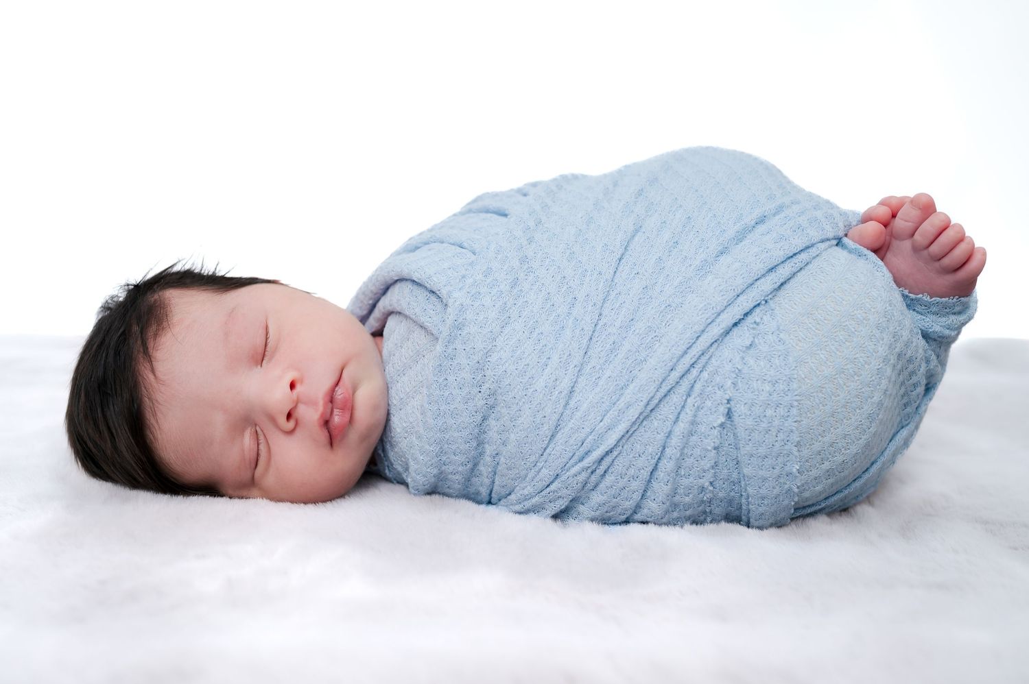 Peaceful newborn baby swaddled in a soft blue wrap, sleeping on a white fluffy blanket.