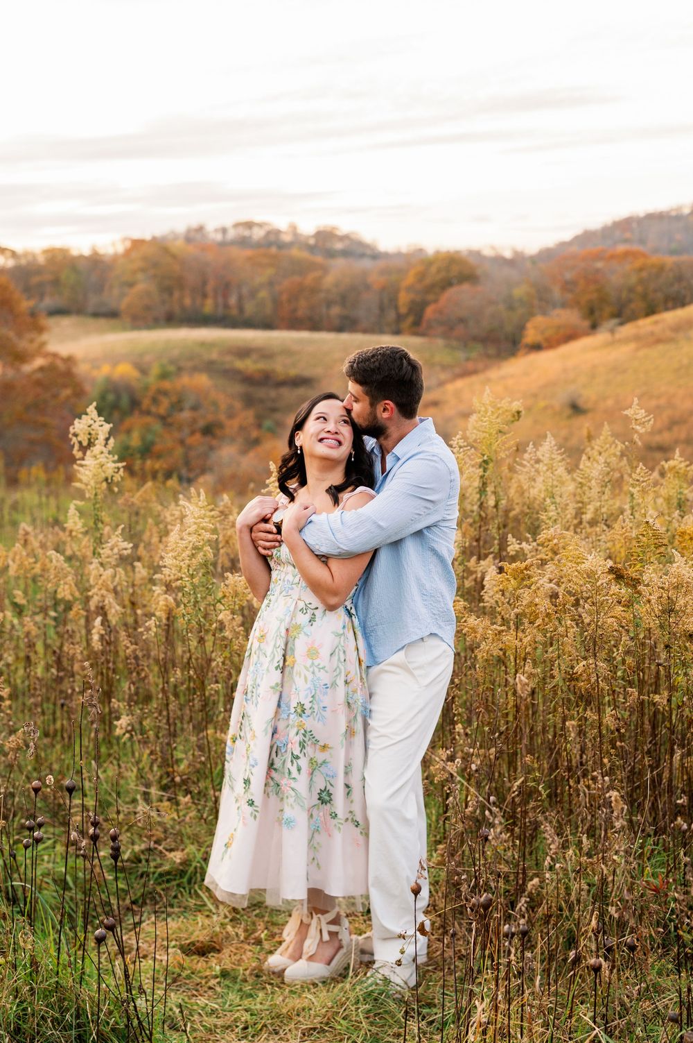 Couple embracing in autumn field, woman in floral dress laughing as man kisses her cheek, fall foliage hills behind.