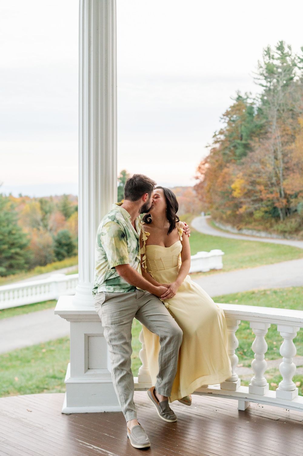Couple sharing a romantic moment on a white porch railing with autumn foliage in the background.
