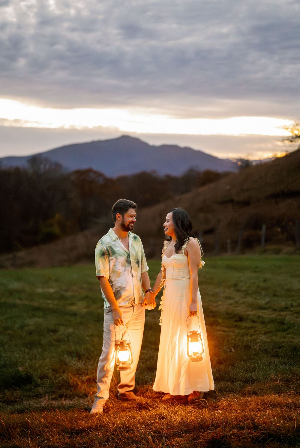 Couple holding glowing lanterns at sunset in a mountain meadow, woman in white dress, man in tie-dye shirt.