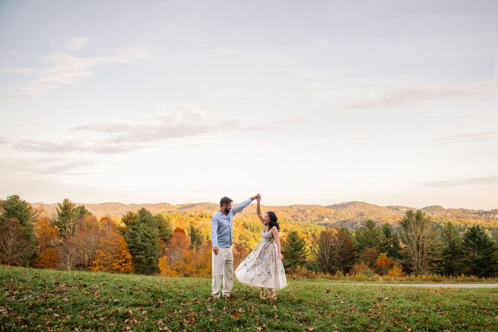 Couple dancing together on a green hillside with vibrant autumn foliage and mountains in the background at sunset.