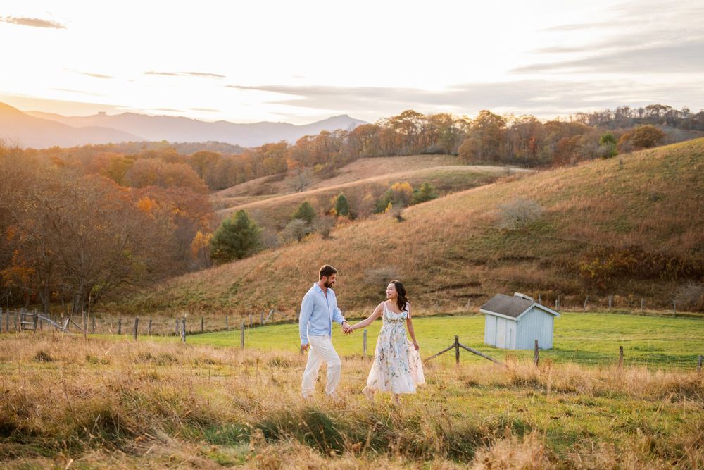 Couple holding hands walking through golden autumn hills at sunset with mountain views and a small barn in background.