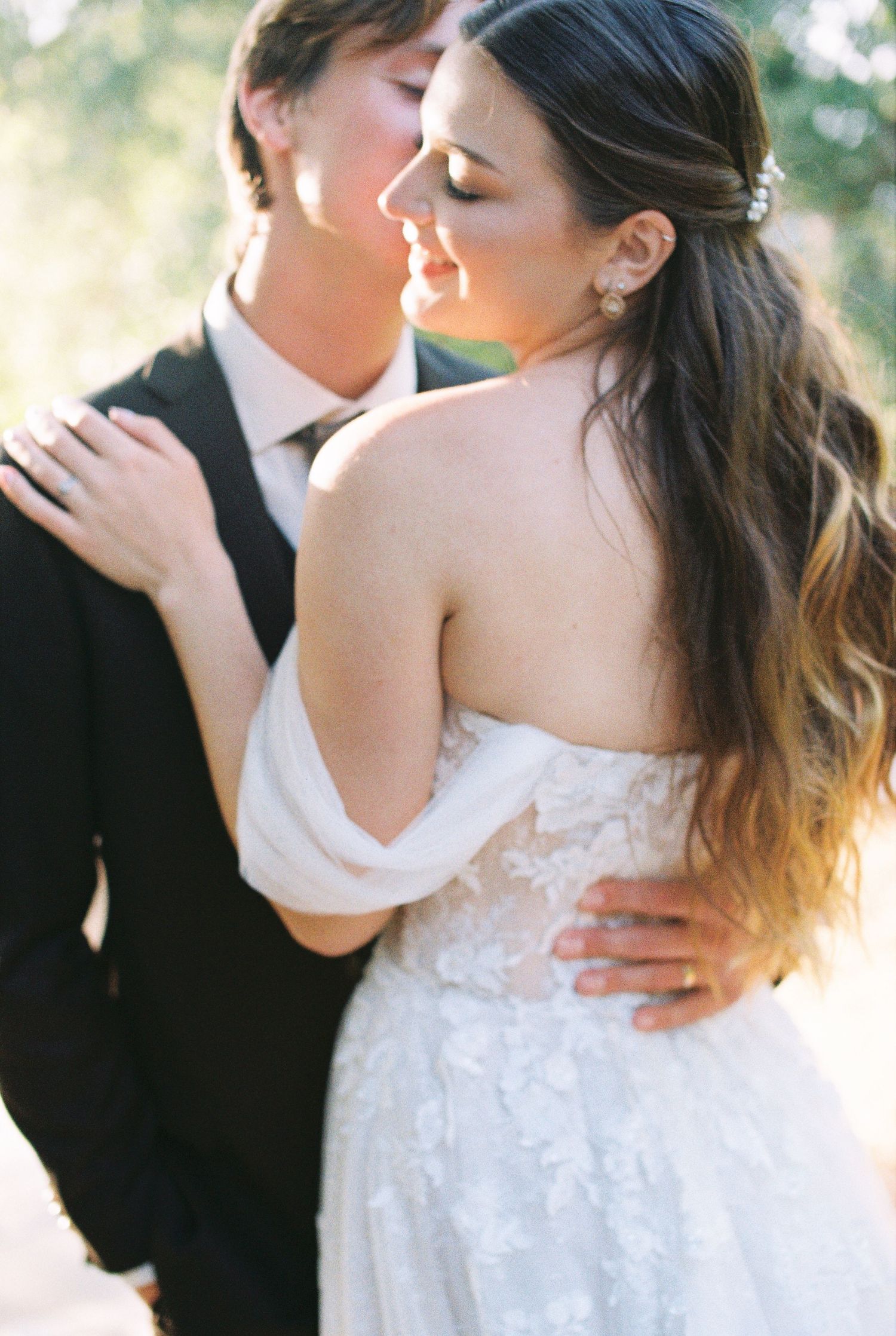 Romantic wedding photo captured through flowing white veil with bride in lace dress and groom in black suit sharing a kiss outdoors