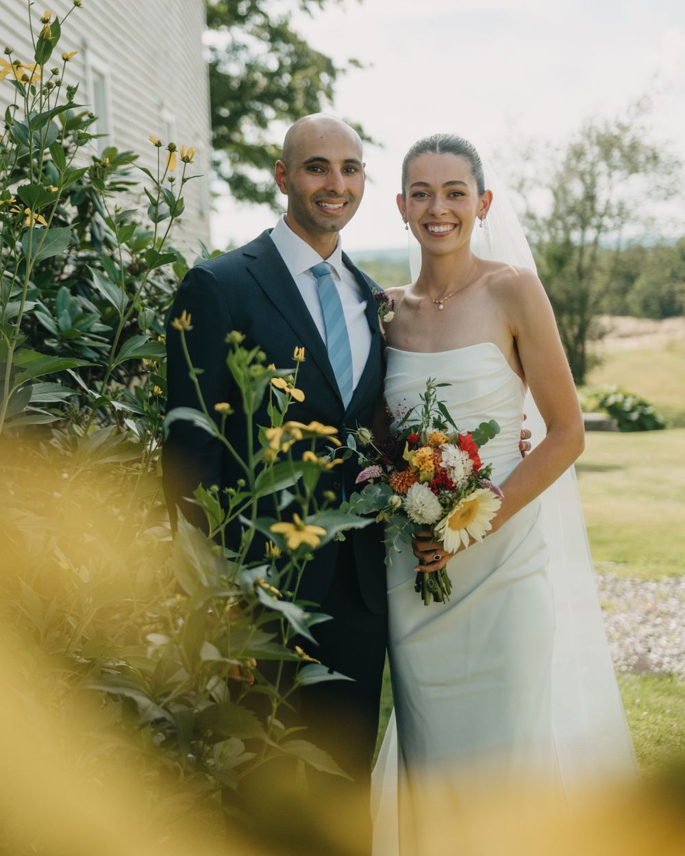 Smiling bride and groom pose together outdoors among yellow wildflowers, bride holding colorful bouquet in white strapless gown.