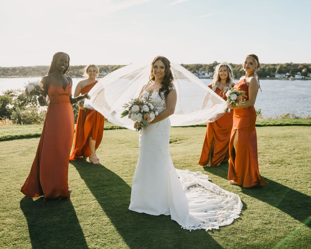 Bride in white gown with flowing veil surrounded by four bridesmaids in burnt orange dresses outdoors.