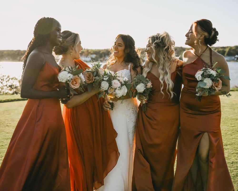 Bride and bridesmaids in rust orange gowns laughing together holding bouquets by waterfront at sunset.