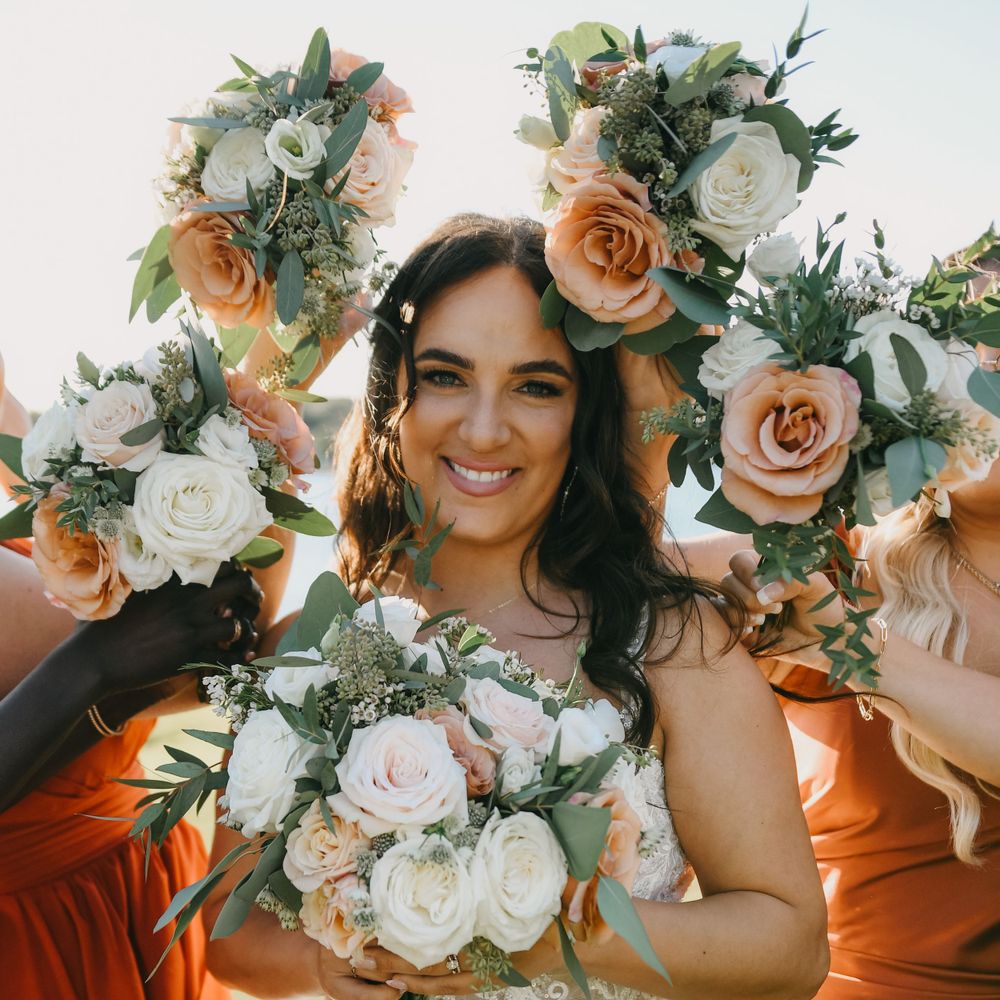 Bride smiling surrounded by bridesmaids holding up peach and white rose bouquets in bright sunlight.