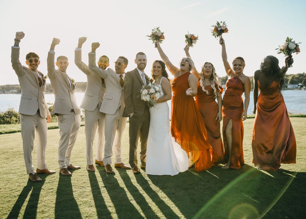Wedding party celebrating outdoors, bridesmaids in rust orange dresses raising bouquets with groomsmen in tan suits by waterfront.