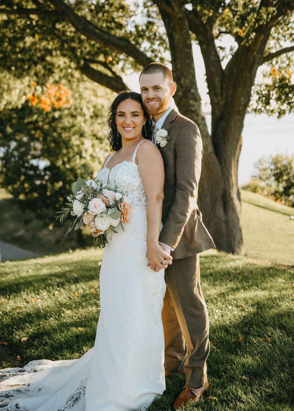 Bride in lace gown holding white bouquet poses with groom in brown suit under large tree at golden hour.