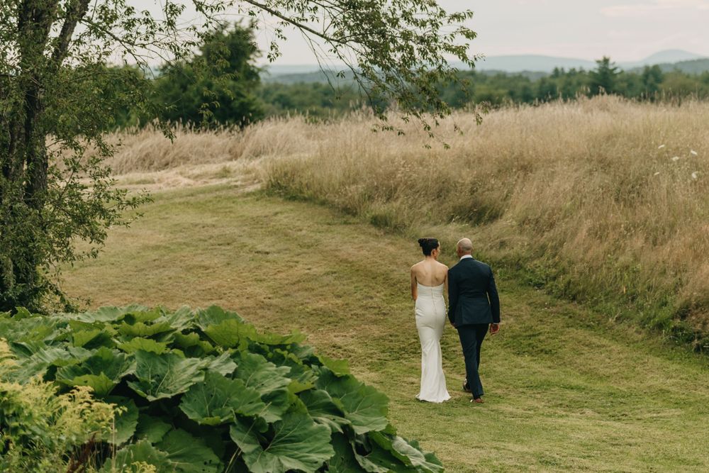 Bride and groom walking hand in hand through lush green meadow with golden fields in background.