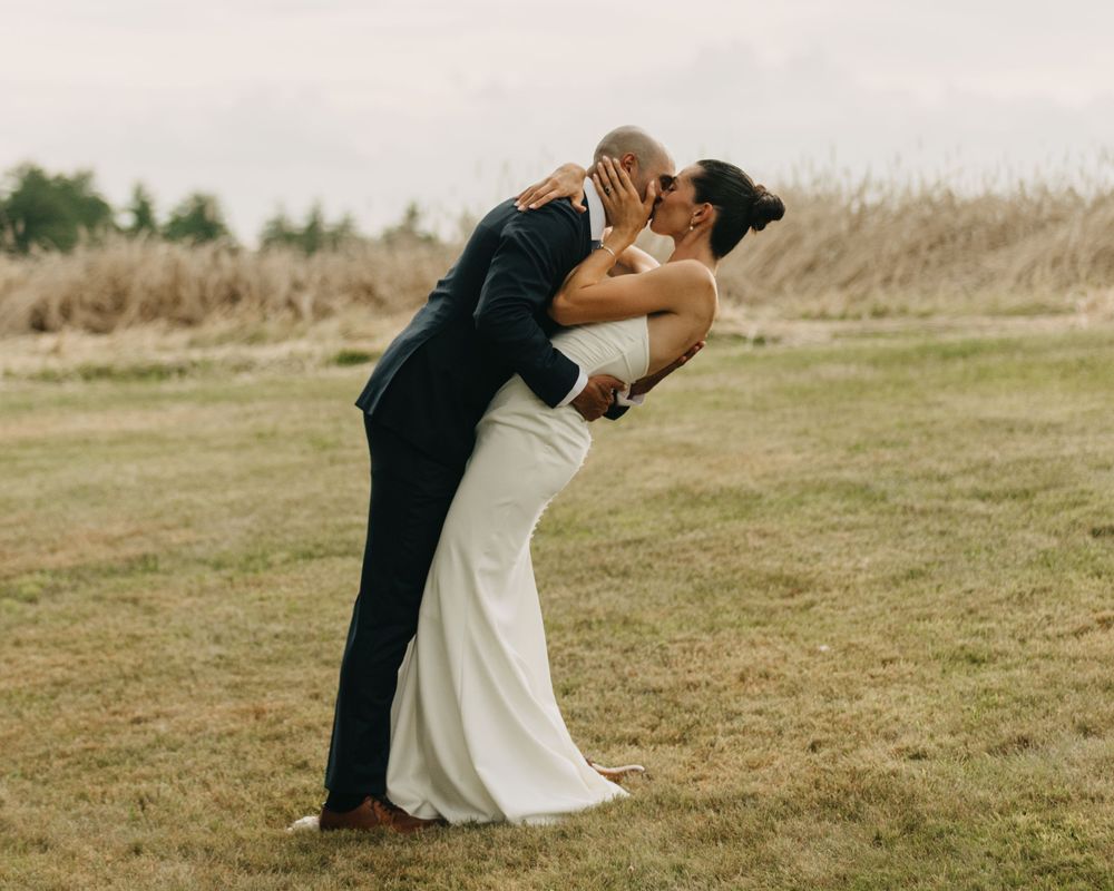 Groom dips bride in white wedding gown for a romantic kiss on a grassy outdoor field.