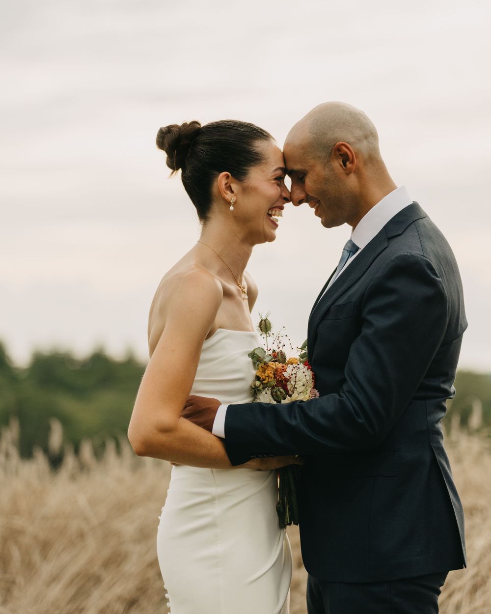 Bride and groom share intimate moment in golden field, touching foreheads while holding bouquet.