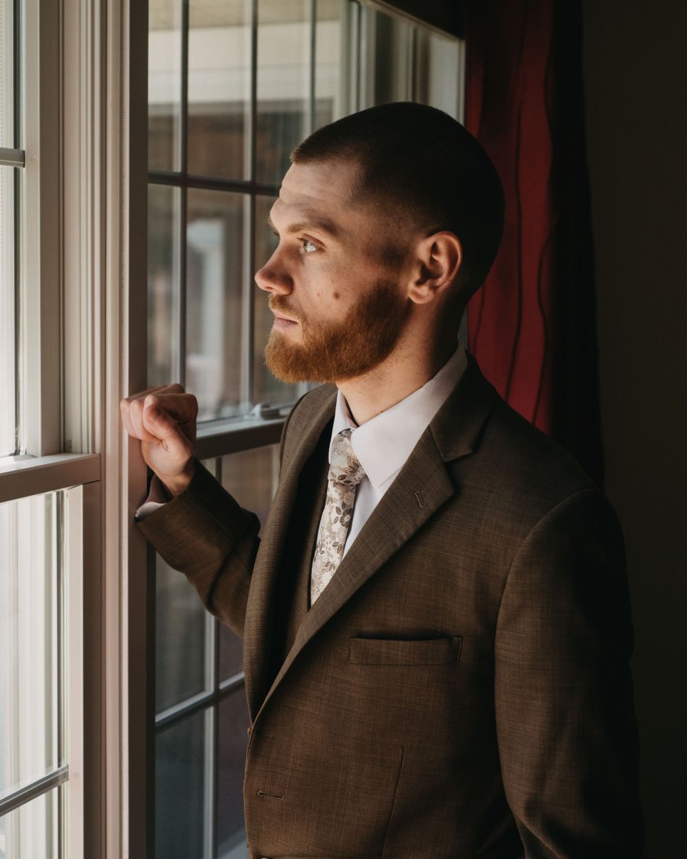 Red-bearded groom in brown suit gazes thoughtfully out window with soft natural light before wedding ceremony.
