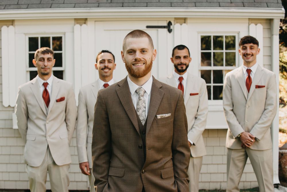 Groom in brown suit stands with four groomsmen in tan suits and red ties outside a white building.