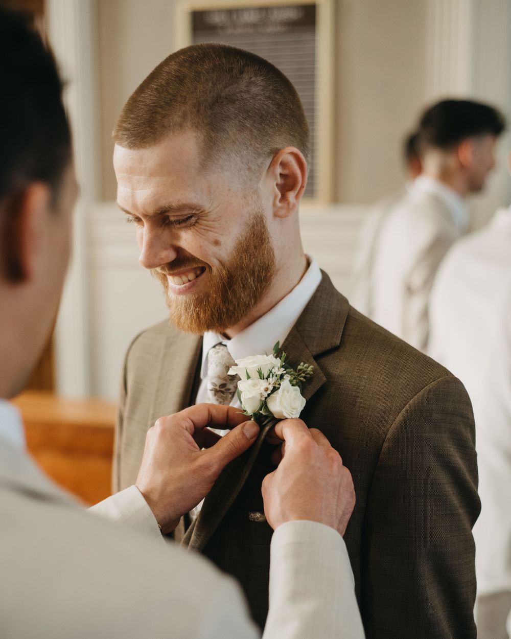 Smiling red-bearded groom in brown suit has white rose boutonniere pinned to lapel before wedding ceremony.