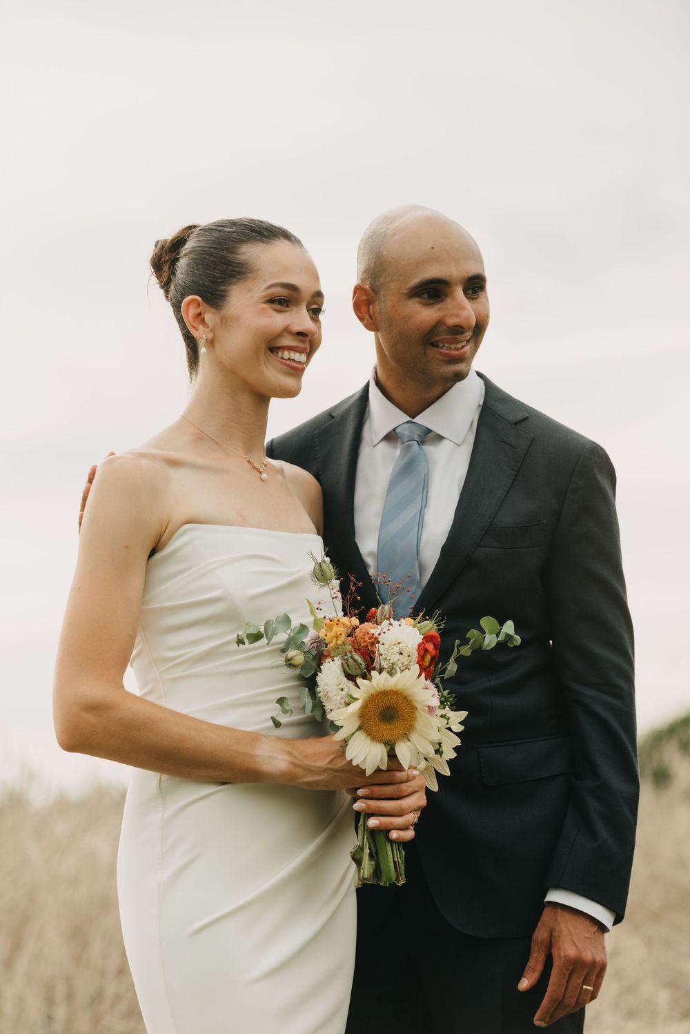 Smiling bride in strapless gown holds sunflower bouquet beside groom in dark suit outdoors.