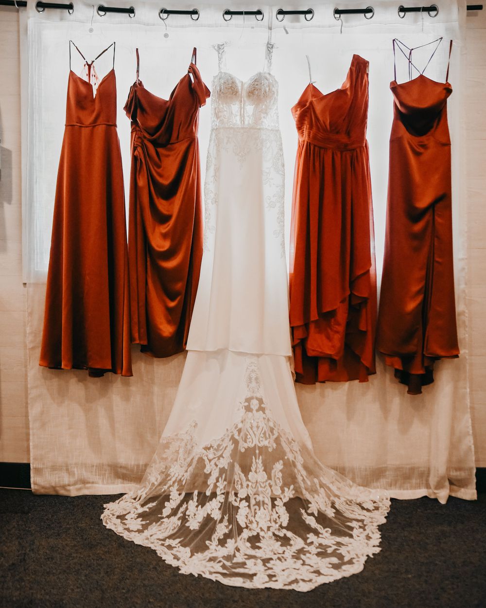 White lace wedding gown displayed alongside four rust-colored satin bridesmaid dresses on hangers.