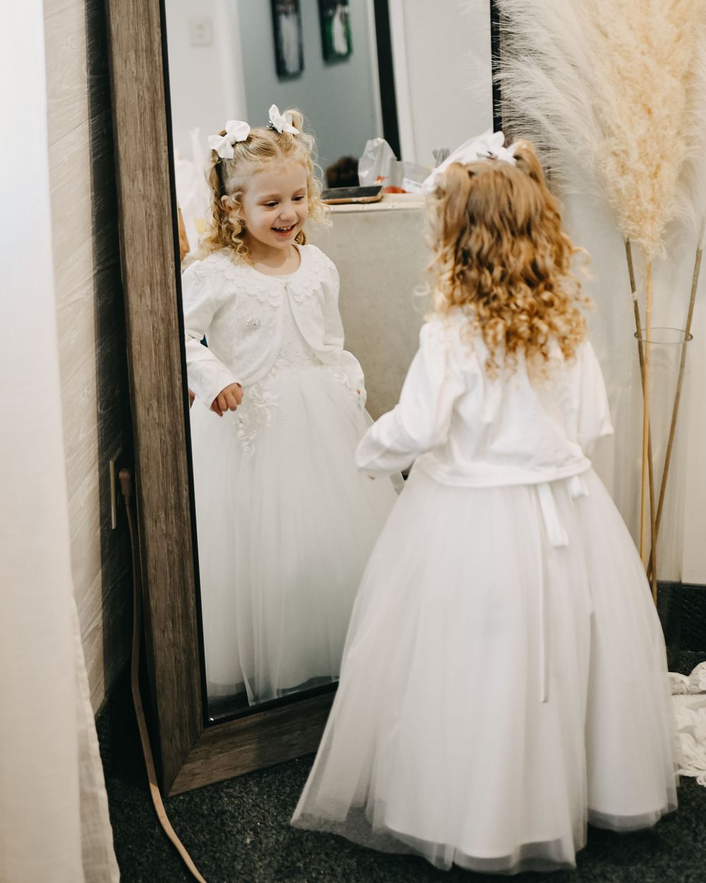 Young flower girl in white dress and bow smiles at her reflection in a large mirror.