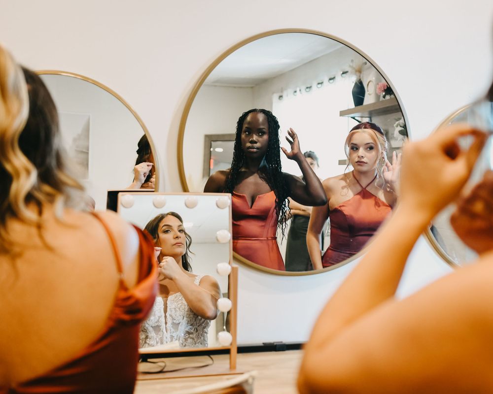 Bridesmaids in rust-colored dresses get ready together reflected in round mirrors.