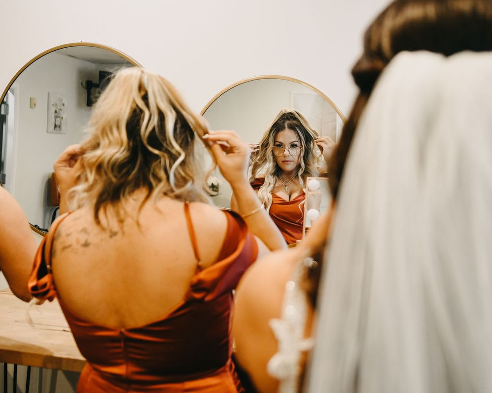 Bridesmaid in rust satin dress helps bride with veil while reflected in a round mirror.