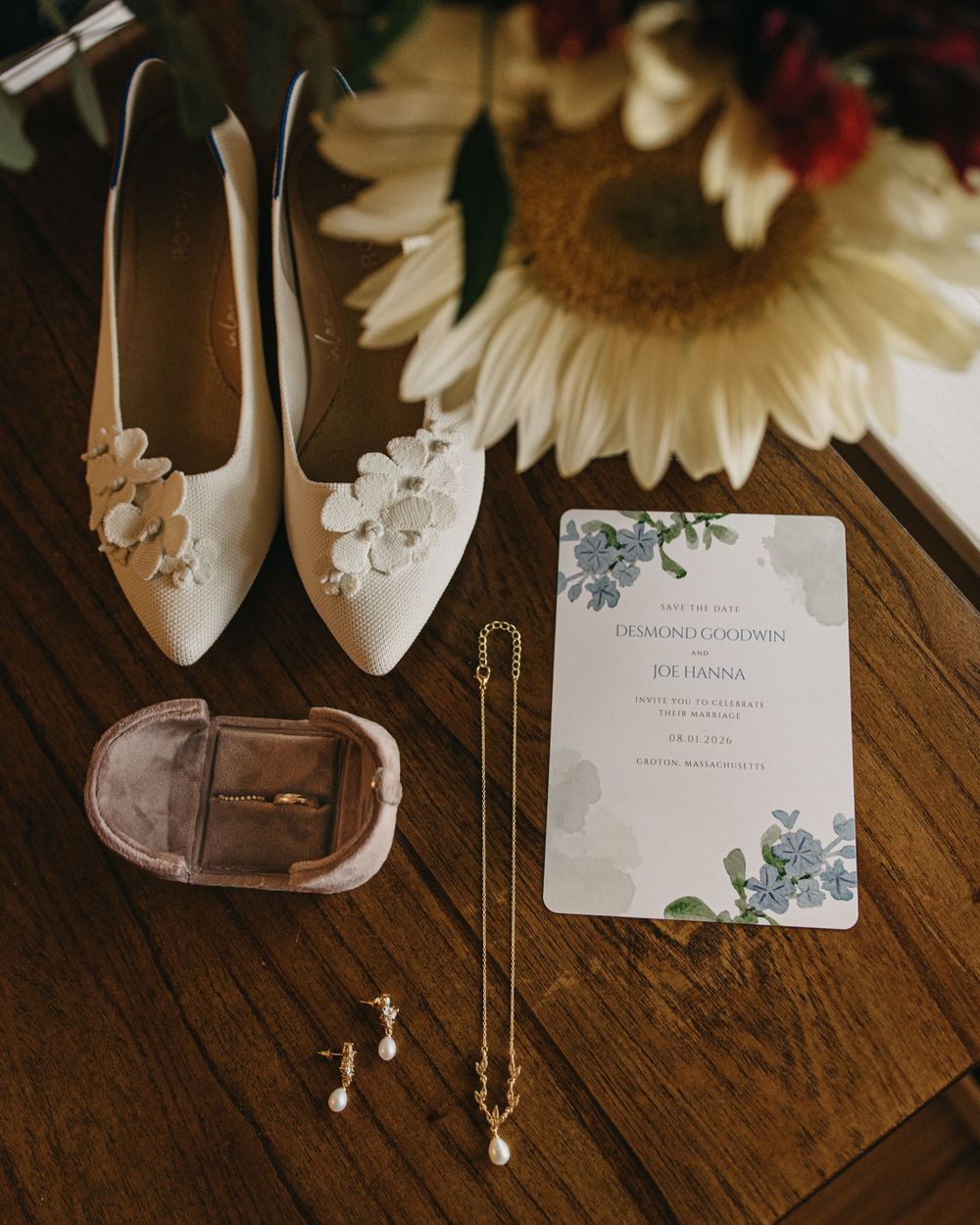 Elegant wedding flat lay featuring floral shoes, ring box, pearl jewelry, and floral invitation beside sunflower.