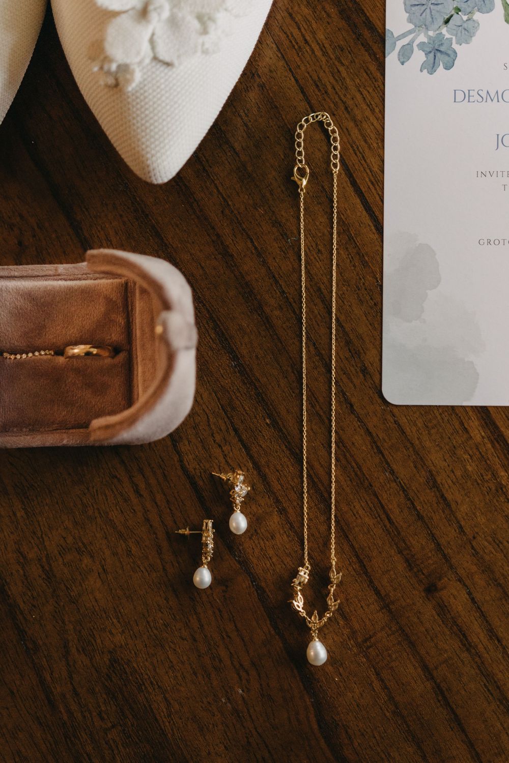 Gold pearl drop necklace and earrings with ring box and wedding invitation on wooden surface.
